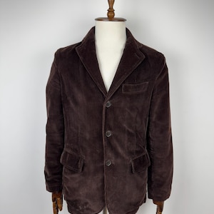 Puede incluir: Una blazer de pana marrón con solapa de muesca, cierre de tres botones y dos bolsillos delanteros. La chaqueta se exhibe en un maniquí de madera. El color de la blazer es un marrón profundo y rico.