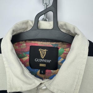 Puede incluir: Una camiseta de rugby a rayas blancas y negras con cuello color crema. Se ve la etiqueta de Guinness, con el logo del arpa y "ESTD 1759". La camiseta est&aacute; colgada en una percha negra.