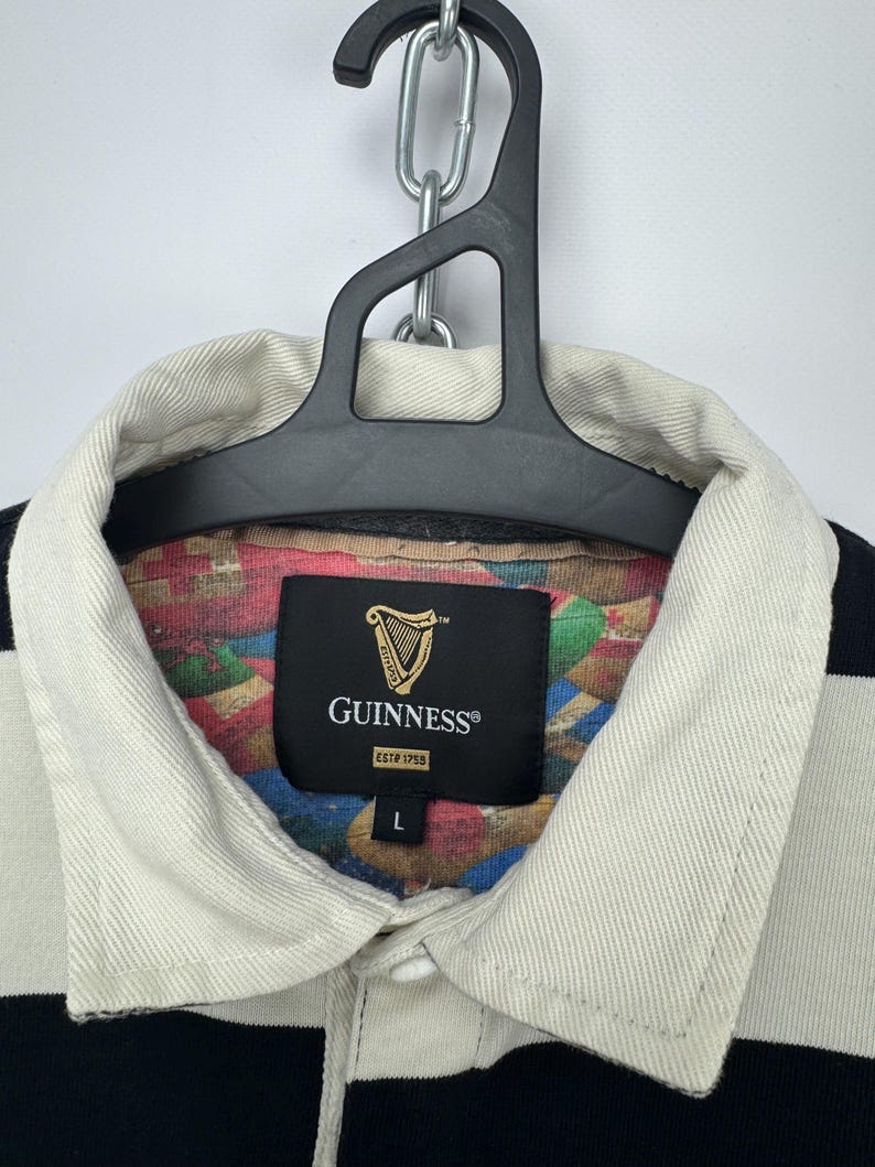 Puede incluir: Una camiseta de rugby a rayas blancas y negras con cuello color crema. Se ve la etiqueta de Guinness, con el logo del arpa y "ESTD 1759". La camiseta est&aacute; colgada en una percha negra.