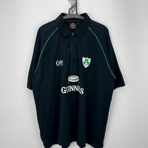 Może przedstawiać: Czarna koszulka polo z zielonymi wykończeniami, białym logo Guinness i zielonym emblematem koniczyny. Zawiera r&oacute;wnież napis "Cotty Trad". Koszulka wisi na metalowym łańcuchu.