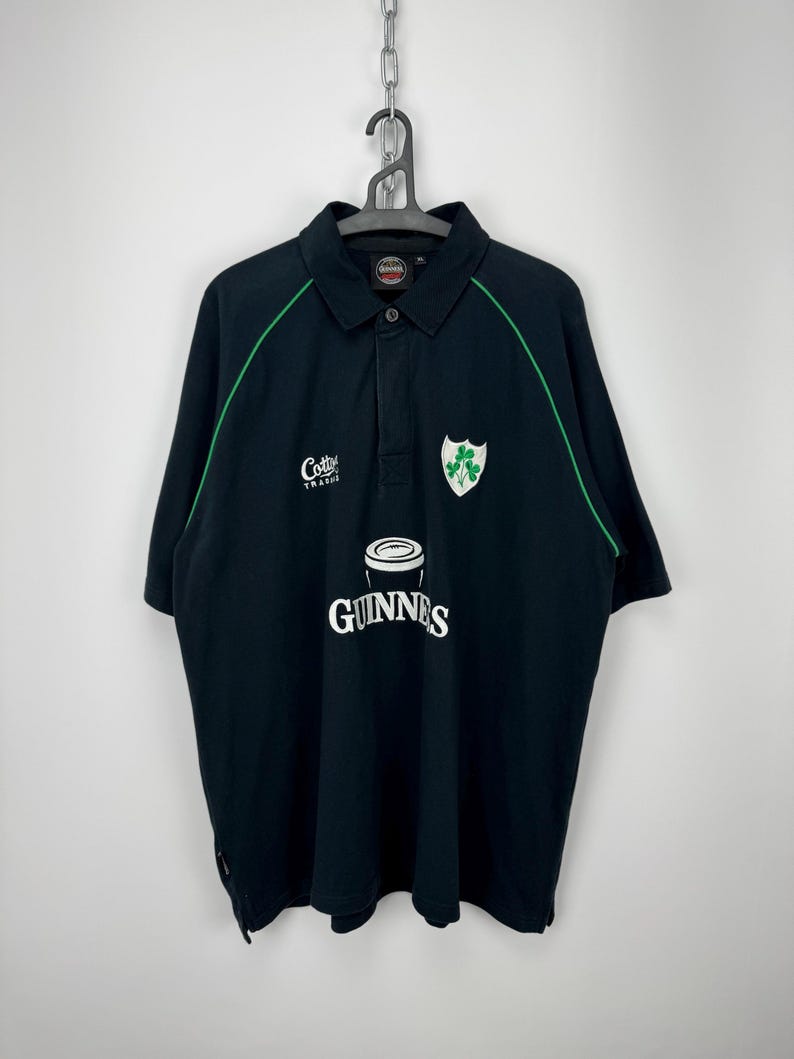 Może przedstawiać: Czarna koszulka polo z zielonymi wykończeniami, białym logo Guinness i zielonym emblematem koniczyny. Zawiera r&oacute;wnież napis "Cotty Trad". Koszulka wisi na metalowym łańcuchu.