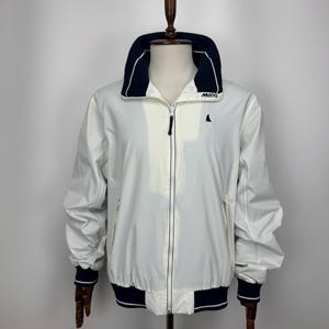 May include: A white zip-up jacket with a navy blue high collar and matching navy blue ribbed cuffs and hem. The jacket features a small black sailboat logo on the left chest and the word MUSTO printed in white on the navy blue collar section.