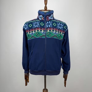 Peut inclure: Veste polaire bleu marine avec un motif géométrique coloré sur la poitrine et les épaules. Le motif comprend des flocons de neige, des losanges et d'autres formes en bleu, vert et rouge. La veste a une fermeture éclair complète et deux poches latérales.