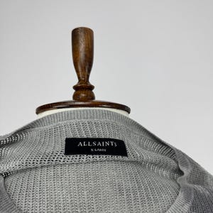 May include: A close-up of a grey, open-knit sweater with the brand name "ALLSAINTS" on a black label. The sweater is size X-LARGE and is displayed on a wooden mannequin.