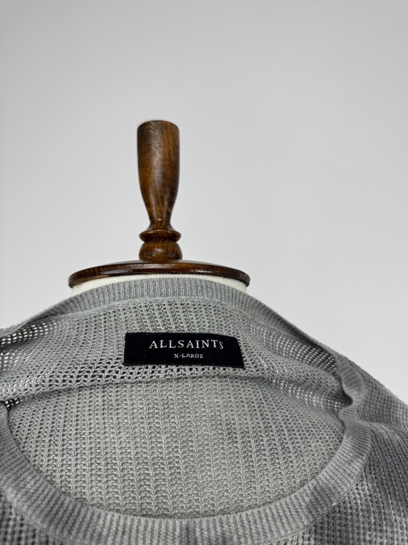 May include: A close-up of a grey, open-knit sweater with the brand name "ALLSAINTS" on a black label. The sweater is size X-LARGE and is displayed on a wooden mannequin.