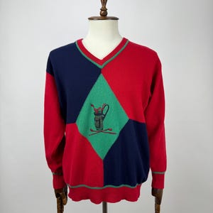 May include: A colour-blocked V-neck sweater in red, navy blue, and green. The sweater features a diamond pattern with an embroidered golf bag and clubs design. The sleeves and sides are red, with navy blue and green diamond shapes on the front.