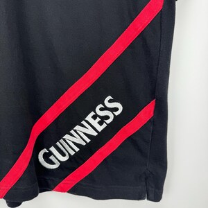Może przedstawiać: Czarny t-shirt z czerwonymi ukośnymi paskami i białym napisem "GUINNESS". Koszulka ma klasyczny kr&oacute;j, z okrągłym dekoltem i kr&oacute;tkimi rękawami. Logo Guinness jest wyraźnie widoczne.