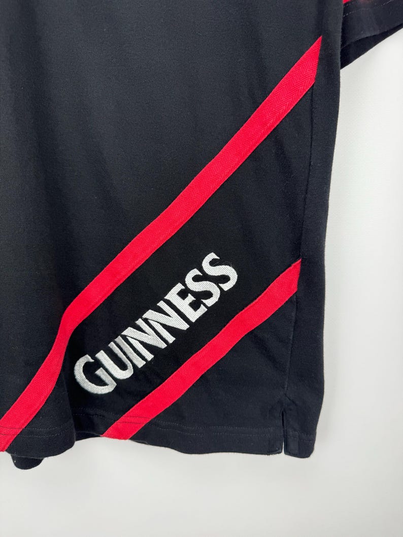 Może przedstawiać: Czarny t-shirt z czerwonymi ukośnymi paskami i białym napisem "GUINNESS". Koszulka ma klasyczny kr&oacute;j, z okrągłym dekoltem i kr&oacute;tkimi rękawami. Logo Guinness jest wyraźnie widoczne.