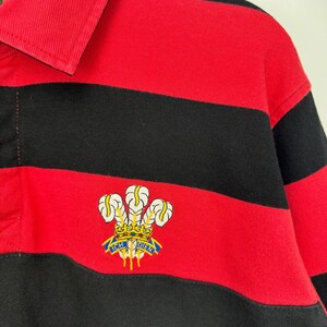Może przedstawiać: Koszulka do rugby w czerwono-czarne poziome paski z kołnierzem. Koszulka posiada haftowany emblemat z napisem "ICH DIEN" i wzorem korony. Koszulka wykonana jest z grubego, wytrzymałego materiału.