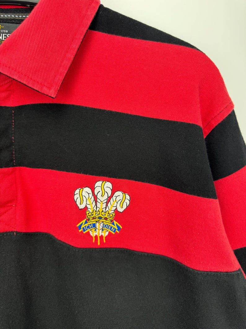 Może przedstawiać: Koszulka do rugby w czerwono-czarne poziome paski z kołnierzem. Koszulka posiada haftowany emblemat z napisem "ICH DIEN" i wzorem korony. Koszulka wykonana jest z grubego, wytrzymałego materiału.