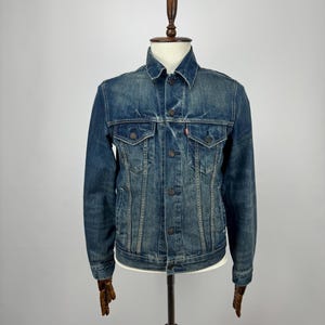 Puede incluir: Chaqueta vaquera azul con un diseño clásico, con cierres de botón, bolsillos en el pecho y cuello. La chaqueta se exhibe en un maniquí de madera, mostrando su ajuste y estilo. El denim tiene un aspecto descolorido y desgastado.