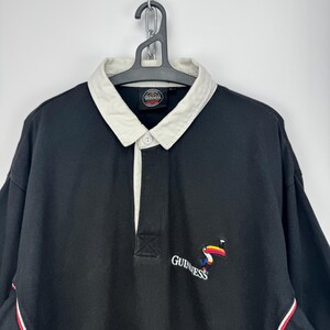 Puede incluir: Camisa negra de manga larga con cuello blanco y tapeta abotonada. La camisa presenta un logotipo de Guinness con un tuc&aacute;n y la marca de palabras Guinness. Rayas rojas y blancas acent&uacute;an las mangas.