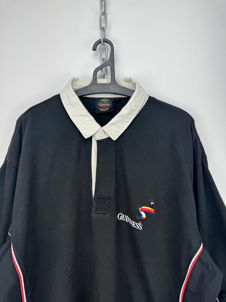 Puede incluir: Camisa negra de manga larga con cuello blanco y tapeta abotonada. La camisa presenta un logotipo de Guinness con un tuc&aacute;n y la marca de palabras Guinness. Rayas rojas y blancas acent&uacute;an las mangas.