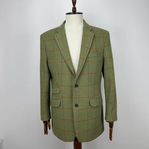 Può includere: Un blazer in tweed verde con motivo a quadri marrone. Il blazer ha due tasche anteriori, una tasca sul petto e una chiusura a due bottoni. È esposto su un manichino di legno.