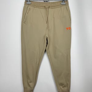 May include: Tan Y-3 sweatpants with a drawstring waist and side pockets. The pants have a ribbed ankle cuff and the Y-3 logo in orange on the right leg. The pants are hanging on a hanger.
