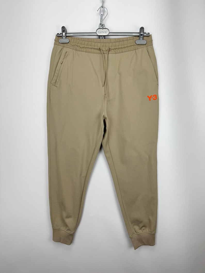 May include: Tan Y-3 sweatpants with a drawstring waist and side pockets. The pants have a ribbed ankle cuff and the Y-3 logo in orange on the right leg. The pants are hanging on a hanger.
