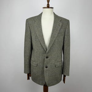 Peut inclure: Un blazer en tweed gris et vert, présenté sur un mannequin en bois. La veste présente une fermeture classique à deux boutons, des revers crantés et deux poches avant. Le tissu présente un motif à carreaux subtil.