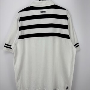 May include: White polo shirt with black horizontal stripes across the upper back and sleeves. The word "GUINNESS" is printed in black on the back collar. The shirt is hanging on a metal hanger.