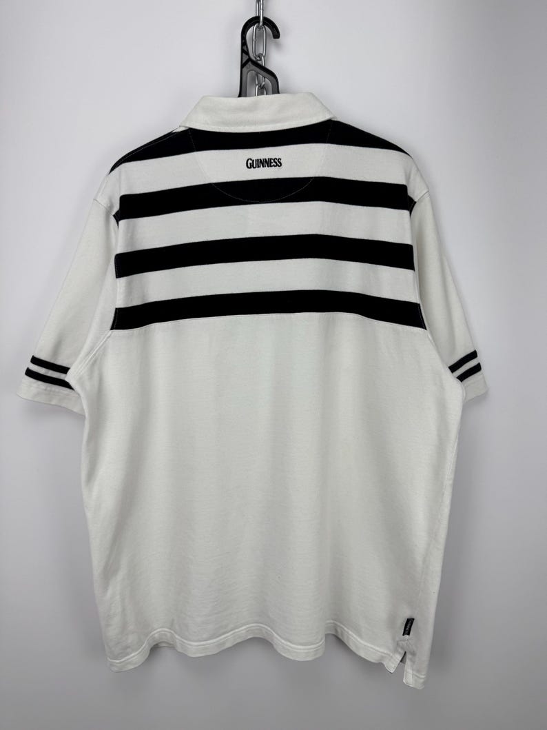 May include: White polo shirt with black horizontal stripes across the upper back and sleeves. The word "GUINNESS" is printed in black on the back collar. The shirt is hanging on a metal hanger.