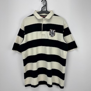 Puede incluir: Polo a rayas crema y negro con mangas cortas. La camisa tiene un peque&ntilde;o emblema con la bandera de la Union Jack en el pecho izquierdo. La camisa est&aacute; colgada de una percha negra.