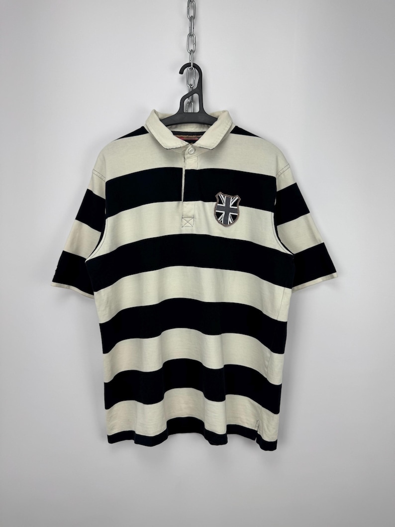Puede incluir: Polo a rayas crema y negro con mangas cortas. La camisa tiene un peque&ntilde;o emblema con la bandera de la Union Jack en el pecho izquierdo. La camisa est&aacute; colgada de una percha negra.