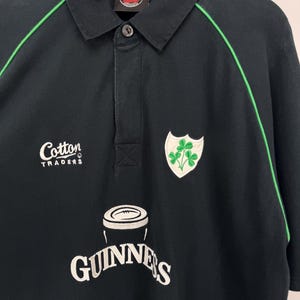 Może przedstawiać: Czarna koszulka polo z zielonymi wykończeniami i kołnierzykiem zapinanym na guziki. Posiada logo Guinness i herb koniczyny. Koszulka ma r&oacute;wnież logo Cotton Traders. Rozmiar XL.