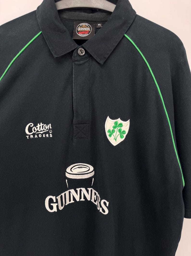 Może przedstawiać: Czarna koszulka polo z zielonymi wykończeniami i kołnierzykiem zapinanym na guziki. Posiada logo Guinness i herb koniczyny. Koszulka ma r&oacute;wnież logo Cotton Traders. Rozmiar XL.