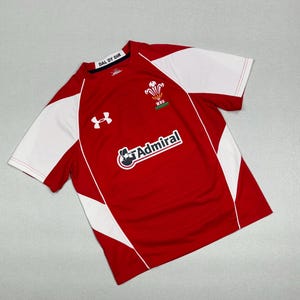 Op de afbeelding: Rood en wit rugby shirt met het Under Armour logo, een Welshe draak en de tekst "WRU" en "Admiral".