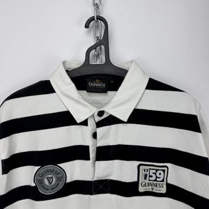 May include: A rugby shirt with black and white horizontal stripes, a collared neckline, and a button placket. The shirt has Guinness patches on the chest. It is hanging on a black hanger. The shirt is a classic style.