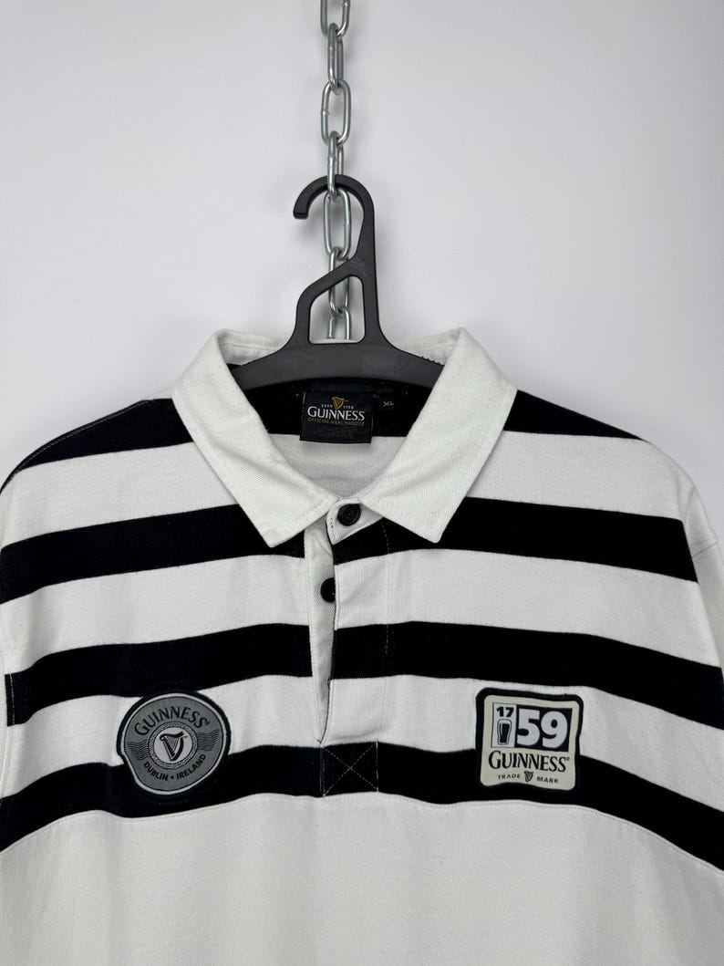 May include: A rugby shirt with black and white horizontal stripes, a collared neckline, and a button placket. The shirt has Guinness patches on the chest. It is hanging on a black hanger. The shirt is a classic style.