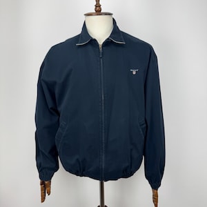 Peut inclure: Une veste zippée bleu marine dotée d'un col classique avec un passepoil clair, de manches longues à poignets élastiques et d'un petit logo Gant brodé sur la poitrine gauche. La veste possède une fermeture éclair intégrale et deux poches latérales.