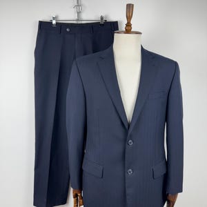 May include: A navy blue suit displayed on a mannequin. The suit comprises a two-button blazer with notched lapels and matching trousers. The trousers are suspended from a metal chain. The suit is made of a textured fabric.