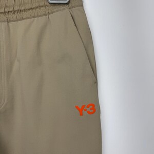 May include: Tan athletic pants with an elastic waistband and a side pocket. The pants feature the orange "Y-3" logo on the leg. The pants are hanging on a black hanger against a white background.