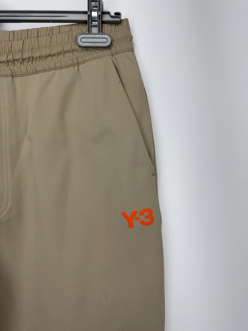 May include: Tan athletic pants with an elastic waistband and a side pocket. The pants feature the orange "Y-3" logo on the leg. The pants are hanging on a black hanger against a white background.