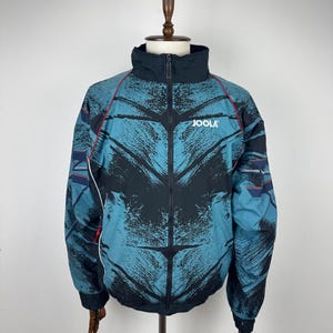 May include: A teal and black athletic jacket with the word "JOOLA" printed on the chest. The jacket has a high collar, a full-length zipper, and red and white accents. The jacket is displayed on a mannequin.