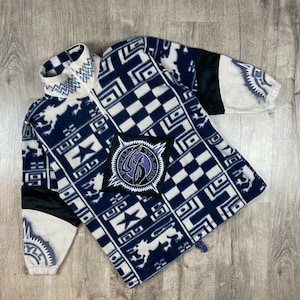Könnte beinhalten: Ein blauer und weißer Fleece-Pullover mit einem schwarz-weißen Karomuster und einem schwarzen Patch mit einem stilisierten Logo. Der Pullover hat einen hohen Kragen und einen Reißverschluss.