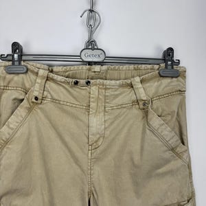 May include: Tan cargo pants with multiple pockets and zippers, hanging on a black hanger. The pants have a relaxed fit and a neutral color, suitable for various outfits. The Getex hanger is attached to a metal chain.