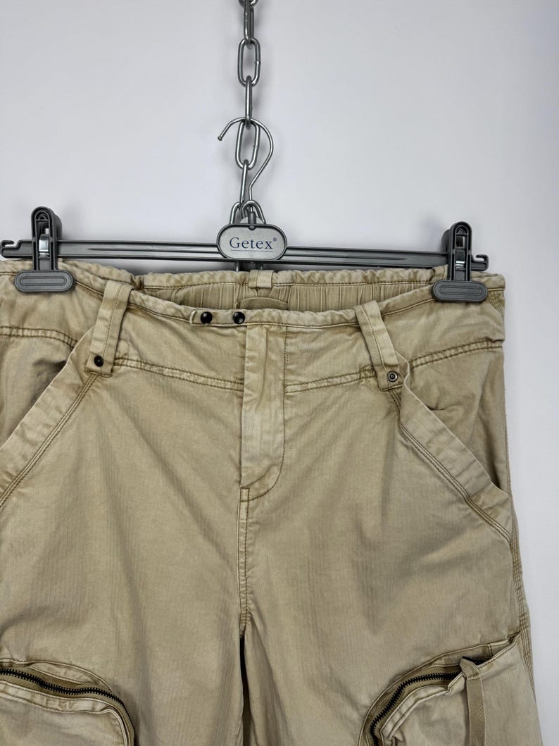 May include: Tan cargo pants with multiple pockets and zippers, hanging on a black hanger. The pants have a relaxed fit and a neutral color, suitable for various outfits. The Getex hanger is attached to a metal chain.