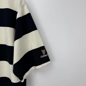 Puede incluir: Camiseta de manga corta a rayas negras y crema. El logotipo de Guinness est&aacute; bordado en la manga izquierda. Las rayas son gruesas y est&aacute;n espaciadas uniformemente. La camiseta es de tejido suave.