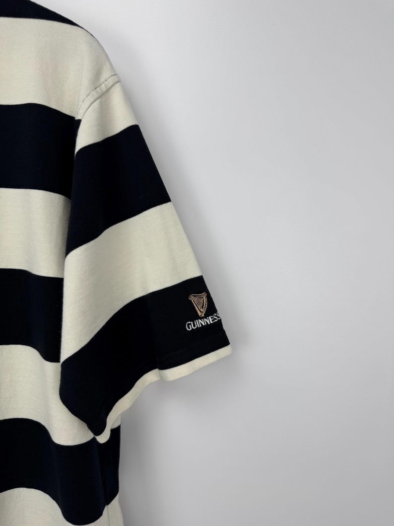 Puede incluir: Camiseta de manga corta a rayas negras y crema. El logotipo de Guinness est&aacute; bordado en la manga izquierda. Las rayas son gruesas y est&aacute;n espaciadas uniformemente. La camiseta es de tejido suave.
