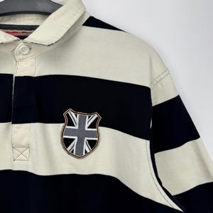 Puede incluir: Polo de manga corta a rayas horizontales en azul marino y crema. La camisa tiene un cuello color crema y un parche con el dise&ntilde;o de la Union Jack en el pecho.
