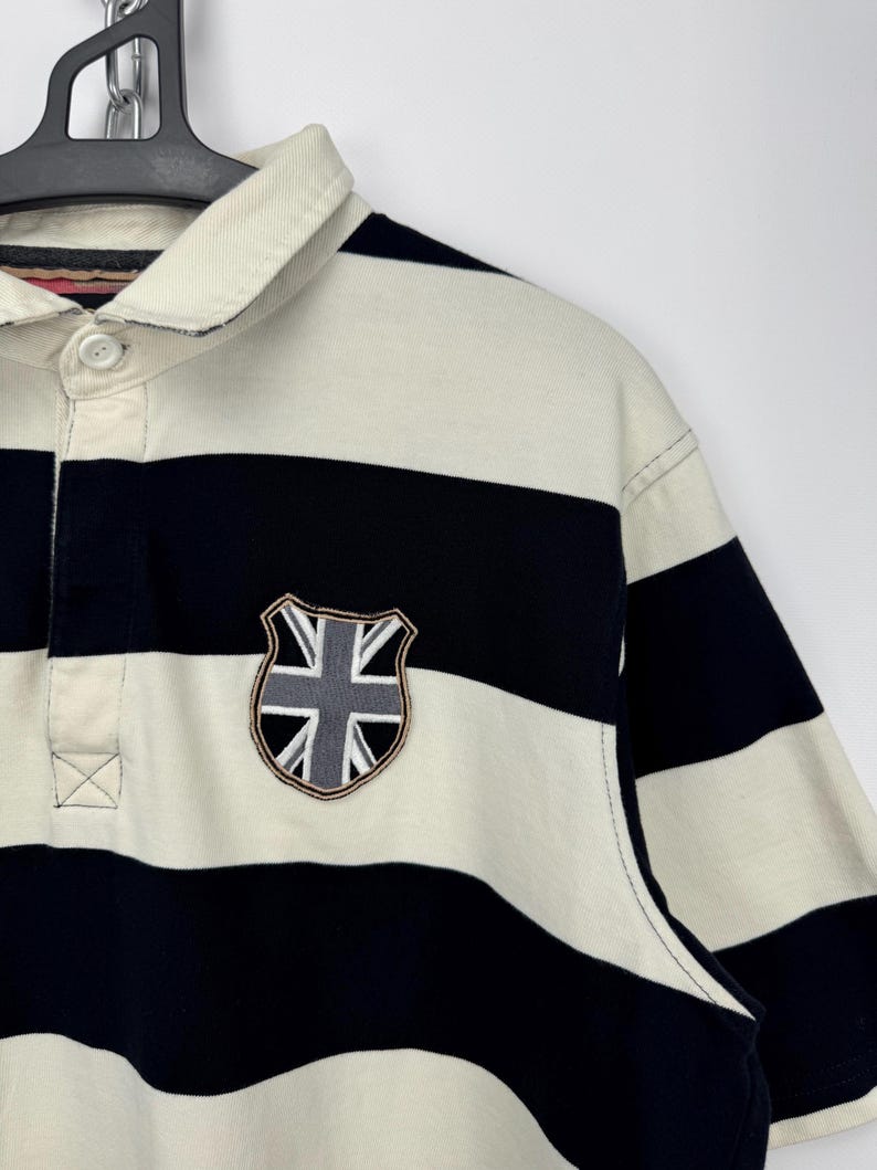 Puede incluir: Polo de manga corta a rayas horizontales en azul marino y crema. La camisa tiene un cuello color crema y un parche con el dise&ntilde;o de la Union Jack en el pecho.