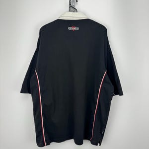 Puede incluir: Polo negro con cuello crema y ribetes rojos y blancos. La palabra "GUINNESS" est&aacute; bordada en rojo en la espalda. La camisa est&aacute; colgada de una percha met&aacute;lica.