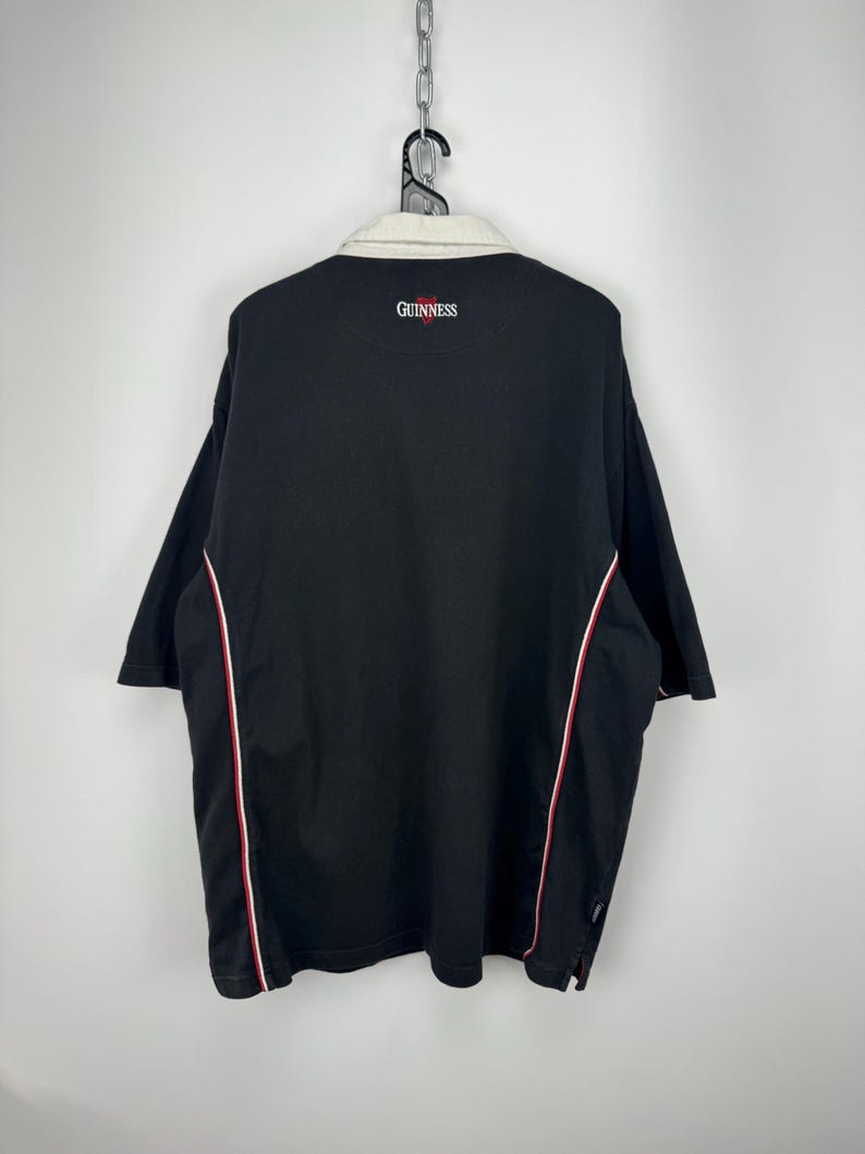 Puede incluir: Polo negro con cuello crema y ribetes rojos y blancos. La palabra "GUINNESS" est&aacute; bordada en rojo en la espalda. La camisa est&aacute; colgada de una percha met&aacute;lica.