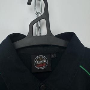Może przedstawiać: Czarna koszula z kołnierzykiem, zielonymi wykończeniami i naszywką z logo Guinness. Koszula wisi na czarnym wieszaku. Etykieta głosi "XL".