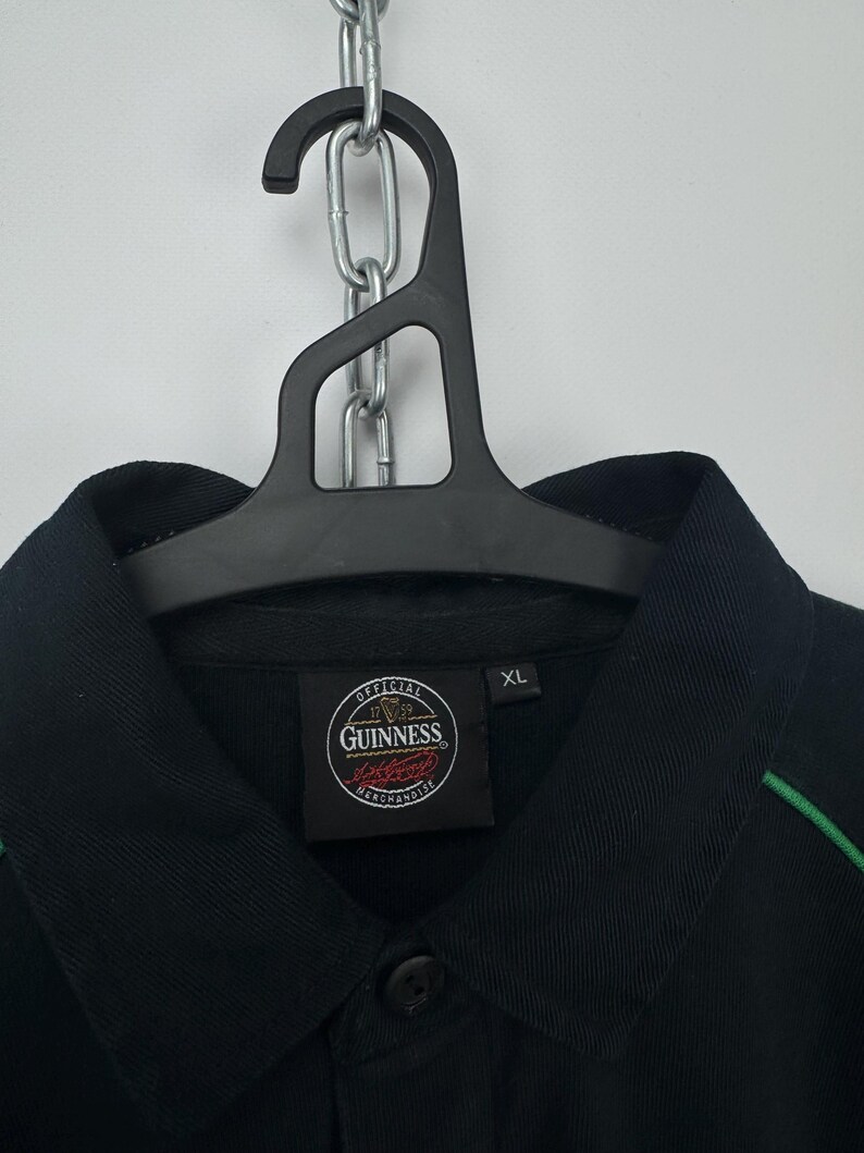 Może przedstawiać: Czarna koszula z kołnierzykiem, zielonymi wykończeniami i naszywką z logo Guinness. Koszula wisi na czarnym wieszaku. Etykieta głosi "XL".