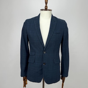 May include: A navy blue blazer with a subtle plaid pattern. The jacket has two front pockets and a two-button closure. The blazer is displayed on a wooden mannequin.