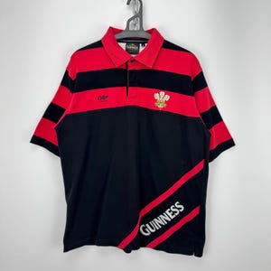 Może przedstawiać: Czarna i czerwona koszulka polo w paski z logo Guinness. Koszulka ma czerwony kołnierzyk i rękawy, z napisem "Cotton" na piersi. Logo Guinness jest wydrukowane w prawym dolnym rogu koszulki.
