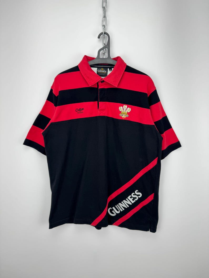 Może przedstawiać: Czarna i czerwona koszulka polo w paski z logo Guinness. Koszulka ma czerwony kołnierzyk i rękawy, z napisem "Cotton" na piersi. Logo Guinness jest wydrukowane w prawym dolnym rogu koszulki.