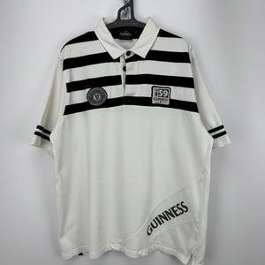 May include: White polo shirt with black stripes across the chest and sleeves. The shirt displays the Guinness logo and the word "Guinness" printed diagonally. The shirt is suspended from a black hanger. The shirt is a classic design.